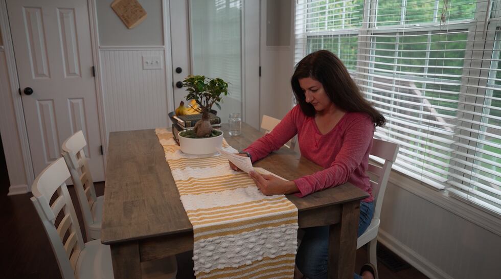 A woman is pictured sitting at a kitchen table, looking down at a piece of paper.
